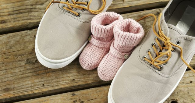 A pair of adult-sized gray sneakers is placed next to tiny pink knitted baby booties on a wooden surface, with copy space. It suggests a heartwarming concept of parenthood and the anticipation of a new family member.