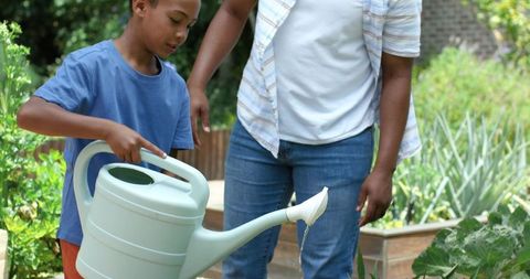 Adult guiding child watering raised garden bed with green watering can, teaching gardening