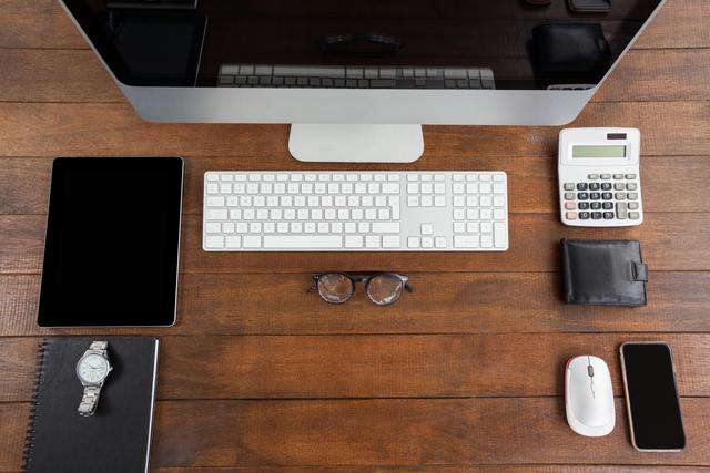 This image shows an overhead view of a neatly organized office desk featuring a computer, digital tablet, mobile phone, keyboard, mouse, calculator, glasses, notebook, watch, and wallet. Ideal for illustrating concepts related to modern workspaces, productivity, business environments, and office organization. Suitable for use in articles, blogs, websites, and marketing materials focused on office setups, technology in the workplace, and efficient work habits.