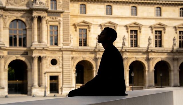Silhouetted man gazing at classical arcade and ornate facade while sitting on stone ledge at golden