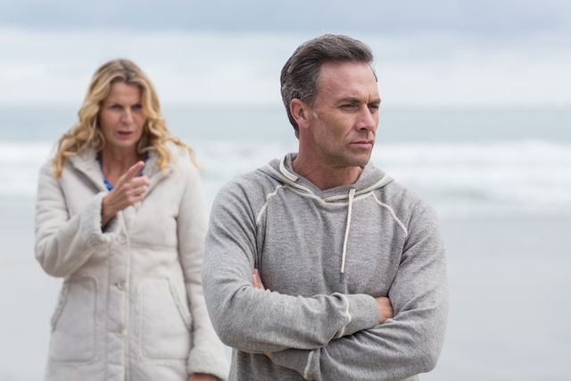 Mature couple having an argument on a beach. The man is standing with arms crossed, looking away, while the woman is gesturing towards him. Both are dressed in winter clothing, indicating a cold day. The ocean and overcast sky create a somber mood. Useful for articles on relationship challenges, communication issues, and emotional conflicts.