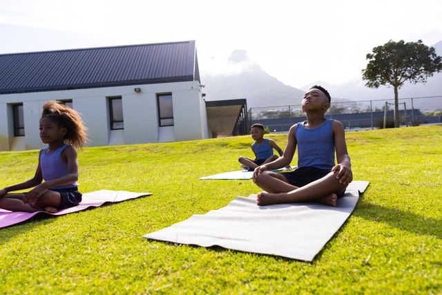Children practicing yoga outdoors at school, sitting on mats on grass. Ideal for promoting physical education, mindfulness, and healthy lifestyle in educational settings. Useful for articles on childhood fitness, school activities, and mental well-being.