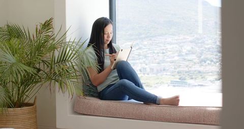 Asian woman sitting on sunlit window seat using stylus on tablet for remote work