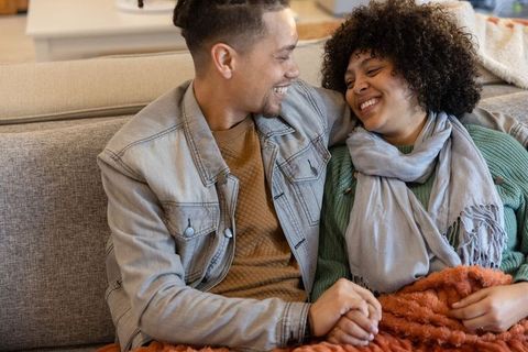 Couple cuddling on sofa holding hands and smiling under cozy orange throw in knitwear