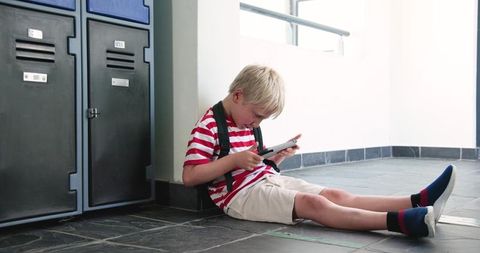 Blond schoolboy sitting on school corridor floor using tablet, wearing backpack, focused