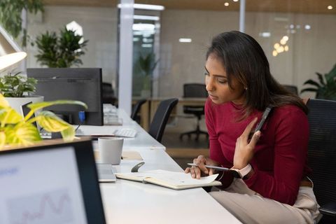 Indian woman writing notes at open-plan office desk holding smartphone and pen