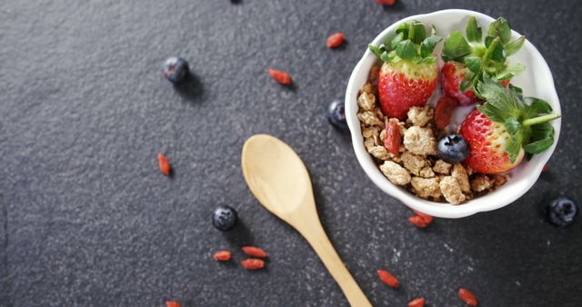 A vibrant, nutritious breakfast bowl featuring fresh strawberries and granola, accompanied by blueberries and goji berries on a dark background, beside a wooden spoon. Perfect for promoting healthy eating, meal planning, and dietary blogs or advertisements.