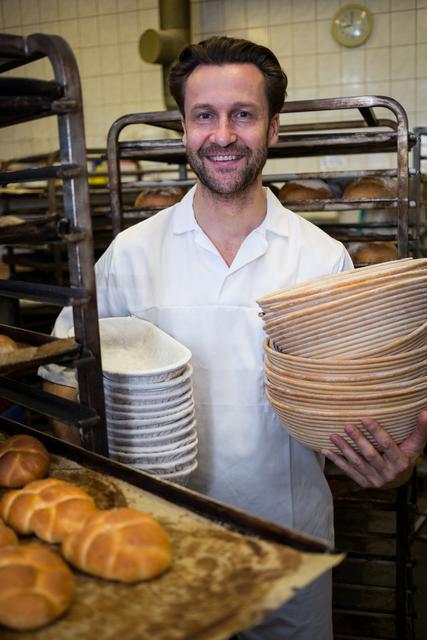 Smiling baker holding trays and baskets in a bakery, surrounded by freshly baked bread. Ideal for use in articles about baking, the food industry, professional chefs, and culinary arts. Perfect for illustrating concepts of happiness at work, artisanal baking, and commercial kitchens.