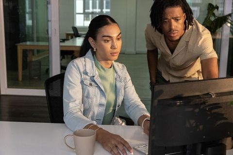 Diverse coworkers collaborating at modern office workstation, woman using monitor and mug