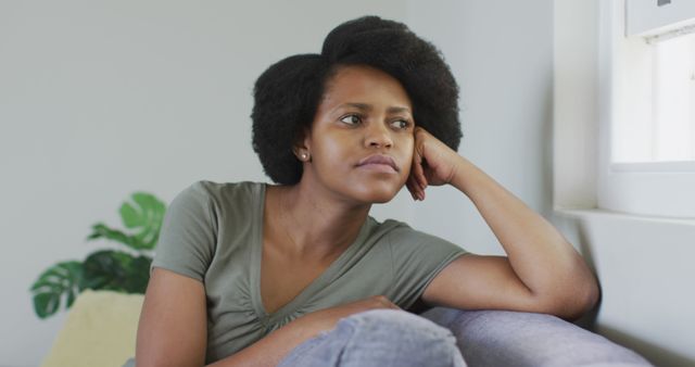 African American woman sitting quietly on couch, leaning her head on hand while looking out window with thoughtful expression. Ideal for use in websites, blogs, articles about mental health, relaxation, urban living, and personal reflections.