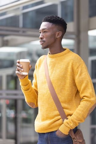 African man holding coffee cup wearing mustard sweater and brown satchel in urban district