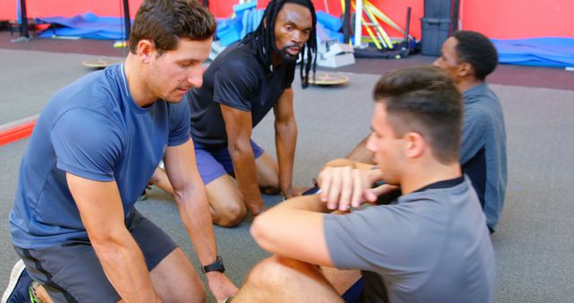 Group of men engaging in physical exercise and training in a fitness center. They are performing sit-ups with two individuals instructing or assisting the others. The indoor gym environment has various equipment in the background, highlighting teamwork and fitness coaching. This image can be used for promoting fitness programs, gym memberships, personal training services, and team-building activities.