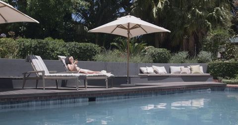 African american woman reclining on poolside lounger in white swimsuit using smartphone