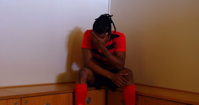 Athlete feeling dejected and disappointed, sitting alone in locker room after a game. Might be used to represent emotions of sadness, defeat, or failure in sports, capturing reflective moments post-competition, or illustrating the impact of loss in athletic contexts.