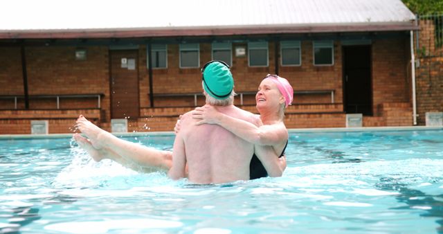 A senior Caucasian woman in a swimming cap is embracing a man in a pool, both smiling and enjoying a leisurely swim. Their joyful interaction suggests a close relationship, sharing a moment of exercise or relaxation together.