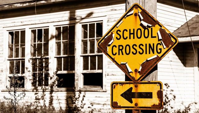 Weathered school crossing sign with peeling paint and rust by abandoned clapboard building