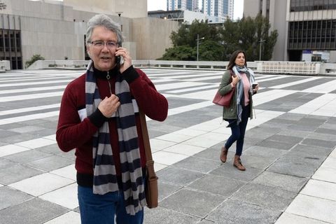 Senior man talking on smartphone while woman walking across modern urban plaza with coffee