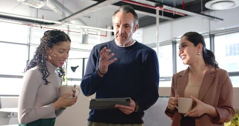 Manager Leading Diverse Team Huddle in Sunlit Open-Plan Office with Tablet and Coffee