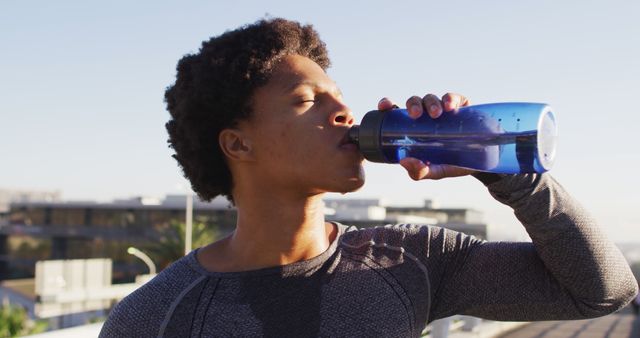 Fit african american man exercising outdoors in city, resting, drinking from water bottle in the sun. fitness and active urban outdoor lifestyle.