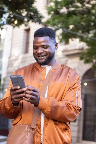 Smiling african man using smartphone wearing rust orange bomber jacket and crossbody bag