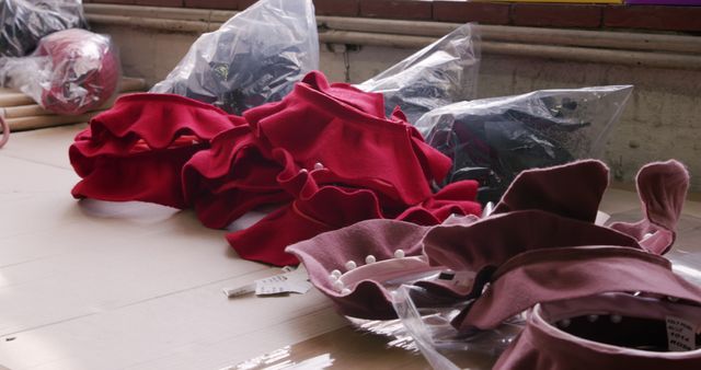 Close-up depicts colorful unfinished garments in sewing workshop on table. Red and purple fabrics with ruffles and parts in clear plastic bags show fashion design and tailoring processes. Ideal for use in articles or content related to fashion industry, textile production, sewing techniques, or creative design studios.