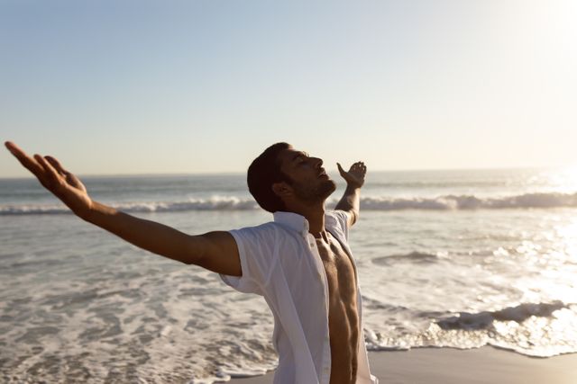 Young man standing with arms outstretched on the beach at sunset, embracing freedom and tranquility. Ideal for use in travel brochures, wellness blogs, lifestyle magazines, and advertisements promoting relaxation and outdoor activities.