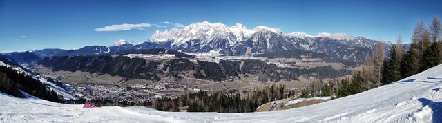 Panoramic view of a snowy mountain range with a valley below, showing forested areas and clear blue skies. Ideal for usage in winter sports advertisements, travel and tourism promotions, outdoor adventure posters, and nature photography galleries.