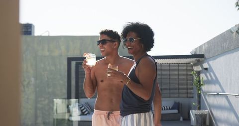 Friends sharing cocktails on rooftop terrace, laughing in sunglasses, summer poolside