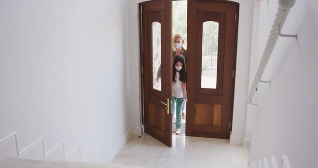 Mother and daughter entering house through an open wooden door while wearing masks. The photo can be used in articles or advertisements related to COVID-19 precautions, family health, or pandemic lifestyle. Excellent for use in health guidelines and social distancing campaigns.