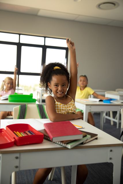 This image depicts a cheerful African American schoolgirl actively participating in a classroom setting by raising her hand. Ideal for educational materials, school brochures, websites focused on childhood education, and articles about student engagement and diversity in schools.