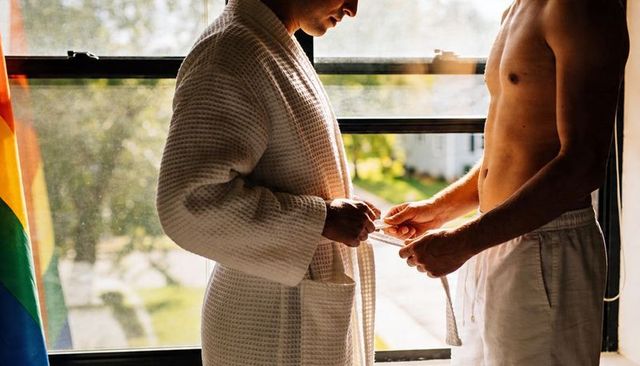 Sunlit intimate male couple tying bathrobe belt by window with pride flag and morning glow