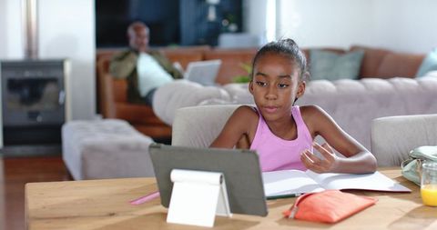 African american girl doing remote learning on tablet at home with dad relaxing