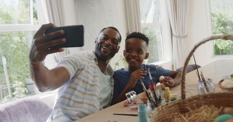 African american father and son taking selfie while painting colorful easter eggs at home