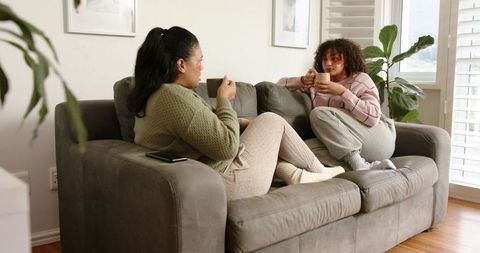 Two Women Relaxing and Chatting on Cozy Gray Sofa With Warm Mugs in Sunlit Living Room
