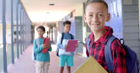 Smiling schoolboy in red plaid shirt holding yellow folder walking locker-lined corridor