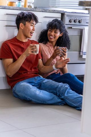 Multiethnic couple sitting on kitchen floor sharing coffee and laughing in cozy home