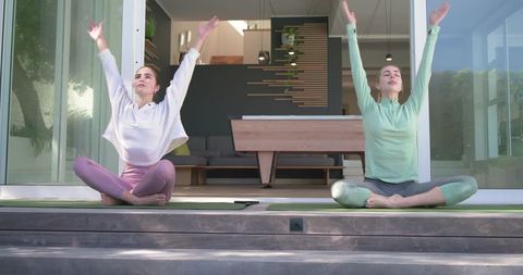 Two women practicing morning yoga and stretching on sunlit modern patio deck