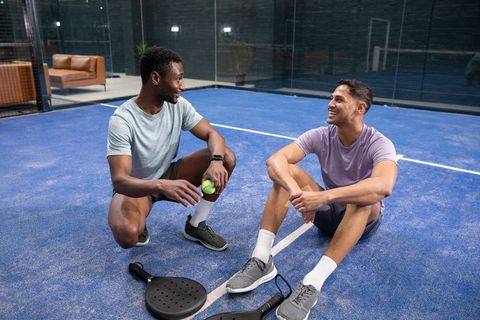 Two friends chatting on blue padel court after match holding ball and rackets