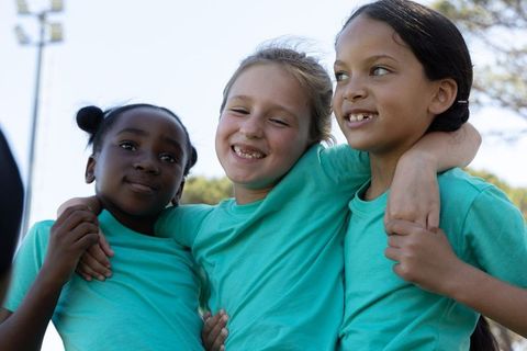 Smiling diverse preteen girls embracing in matching teal shirts on outdoor sports field