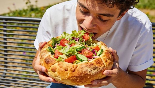 Young man eating giant rustic bread bowl salad on park bench healthy outdoor picnic lunch