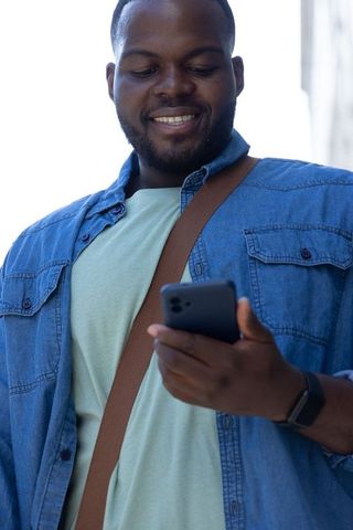 African american man smiling checking smartphone outdoors in denim shirt and crossbody bag