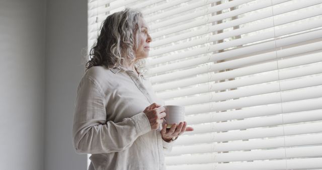 Senior woman with gray hair holding a cup of coffee or tea, standing by a window with blinds. She is wearing casual clothes and looking contemplatively outside. Ideal for themes of solitude, contemplation, peaceful moments, or senior lifestyle. Perfect for articles on aging, health, relaxation at home, and mental wellbeing.