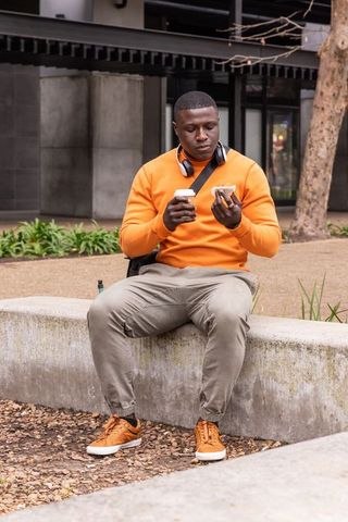 African american man in orange sweater enjoying coffee and sandwich on urban bench