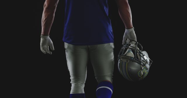 A muscular American football player stands against a dark background, holding a helmet by his side. This image is ideal for promoting sports gear, athletic wear, football events, or anything related to professional athletics. It can also be used in designs for posters, advertisements, and sports magazines.