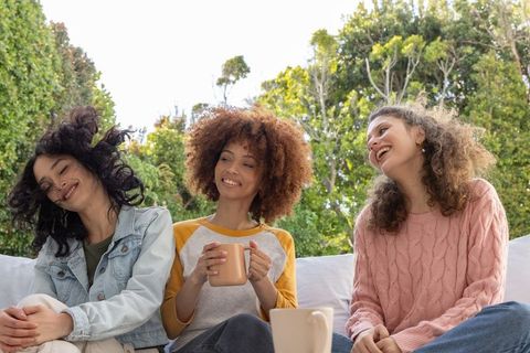 Three friends laughing and holding coffee mugs on backyard sofa, casual outdoor gathering