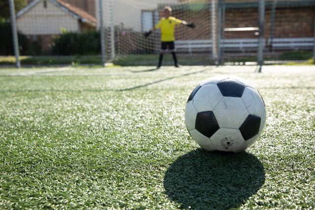 Close up of a football with boy soccer goalkeeper standing in a goal on a green football pitch on a sunny day, waiting with arms out. Childhood healthy lifestyle competition.