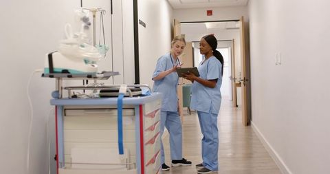 Diverse female nurses reviewing tablet beside crash cart in bright hospital corridor