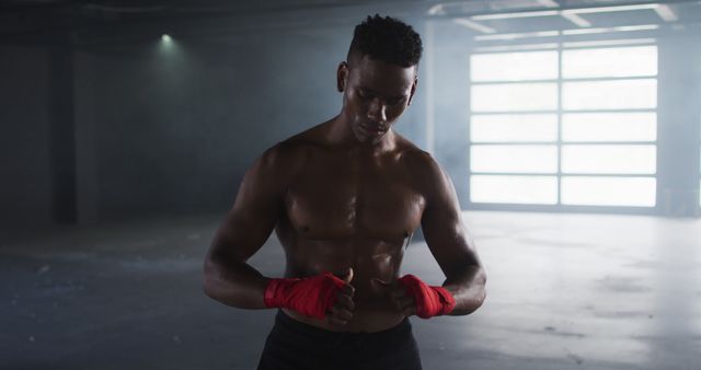 Boxer standing in an industrial gym, wrapping hands with red hand wraps, preparing for training session. Background shows large windows with diffused light and a spacious workout area. Ideal for use in fitness blogs, sports advertisements, and content promoting a healthy lifestyle and physical fitness.