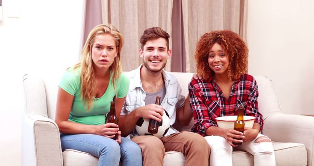 Group of three friends sitting on a couch enjoying a sports game on TV. They are holding beers and snacks like popcorn. The setting is relaxed and homey, indicating a casual gathering. Ideal for illustrating themes of friendship, leisure activities, sports events, and home entertainment. Perfect for use in advertisements, blog posts, social media content, and articles about social gatherings, sports fandom, and young adult lifestyle.