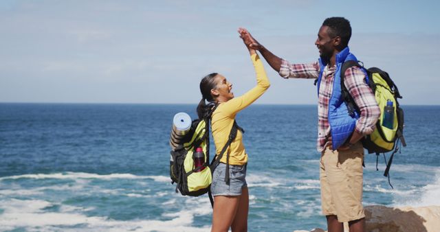 Two hikers are celebrating with a high five on a scenic coastline. They are wearing backpacks, indicating they have been on an adventure. Majestic waves and the ocean are seen in the background. This image could be used for promoting tourism, travel adventures, active lifestyle blogs, and celebrating team achievements in advertisements.